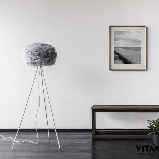 Lampa podłogowa Eos Light z szarym abażurem z piór na białym trójnożnym stojaku, ustawiona w minimalistycznym wnętrzu z białą ścianą, czarno-białą fotografią w ramce i drewnianą ławką z małą rośliną.