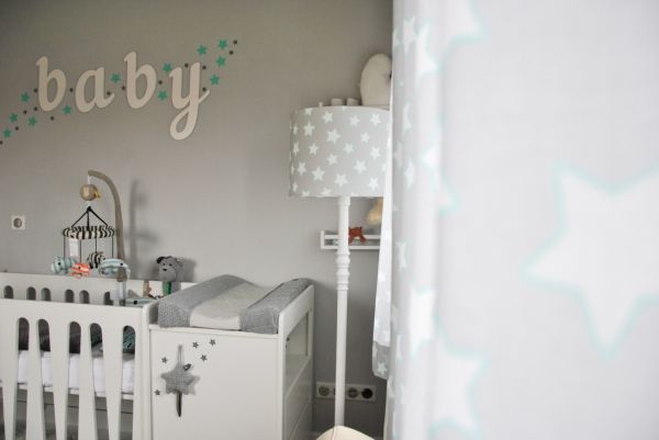 Dziecięca lampa podłogowa Grey Stars z abażurem w biało-szary wzór gwiazdek, ustawiona w jasnym pokoju niemowlęcym obok przewijaka i białej komody, uzupełniająca wystrój z napisem "baby"na ścianie i pastelowymi dekoracjami.