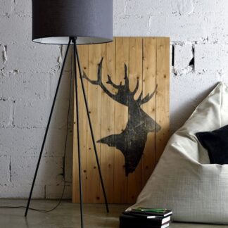 Szara materiałowa lampa podłogowa Simple na czarnym trójnożnym stelażu z tkaninowym abażurem, umieszczona w industrialnym wnętrzu z białą ceglaną ścianą, drewnianą dekoracją z motywem jelenia, jasnymi meblami wypoczynkowymi i betonową podłogą.