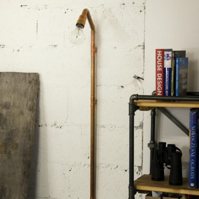 Miedziana lampa ścienna Wand wykonana z rurek hydraulicznych z żarówką Edison. Industrialny design na białej ścianie obok metalowego regału. Minimalistyczny, loftowy styl idealny do surowych, kreatywnych przestrzeni.