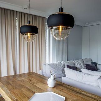 Bursztynowe lampy wiszące Ariel Wide nad drewnianym stołem w jadalni. Nowoczesne oświetlenie z czarnym, płaskim kloszem i szklaną, przezroczystą kopułą. Stylowe lampy sufitowe do kuchni w jasnym wnętrzu z szarą sofą.