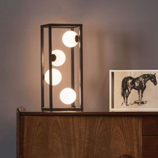Lampa stołowa Ensio z czarną metalową ramą w kształcie prostokątnego prostopadłościanu i czterema kulistymi źródłami światła. Umieszczona na drewnianej komodzie w stylu vintage, obok czarno-białej grafiki przedstawiającej konia. Lampa emituje ciepłe światło, tworząc przytulną atmosferę w nowoczesnym salonie o szarych ścianach.