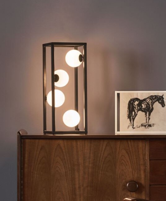 Lampa stołowa Ensio z czarną metalową ramą w kształcie prostokątnego prostopadłościanu i czterema kulistymi źródłami światła. Umieszczona na drewnianej komodzie w stylu vintage, obok czarno-białej grafiki przedstawiającej konia. Lampa emituje ciepłe światło, tworząc przytulną atmosferę w nowoczesnym salonie o szarych ścianach.