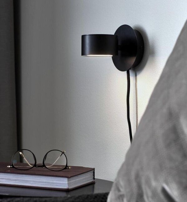 Czarny kinkiet Clyde z okrągłą rozetą i regulowanym, płaskim kloszem na ramieniu. Minimalistyczna lampa do czytania umieszczona przy łóżku z szarą pościelą, obok stolika z książką i okularami. Elegancki, nowoczesny design do sypialni.