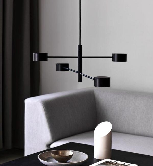 Czarna modernistyczna lampa wisząca Clyde z czterema obrotowymi, cylindrycznymi reflektorami LED na krzyżujących się ramionach, zawieszona nad ciemnym stolikiem kawowym z szarą sofą w tle.