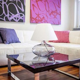 Szklana lampa stołowa Bergen z przezroczystą, fioletową podstawą i białym, stożkowym abażurem na chromowanej nóżce. Ustawiona na nowoczesnym stoliku kawowym z ciemnym blatem w salonie z białą sofą i kolorowymi obrazami w odcieniach fioletu i różu.