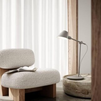 Szara lampa stołowa Stay Long z regulowanym ramieniem i małym, stożkowym kloszem ustawiona na okrągłym, kamiennym stoliku. Minimalistyczny design w neutralnej kolorystyce idealnie komponuje się z kremowym, wełnianym fotelem na drewnianych nogach. Jasne wnętrze z białymi zasłonami i betonową podłogą tworzy spokojną, organiczną przestrzeń do czytania w stylu skandynawskim lub japandi.
