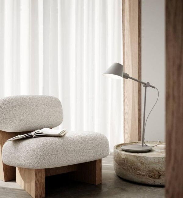 Szara lampa stołowa Stay Long z regulowanym ramieniem i małym, stożkowym kloszem ustawiona na okrągłym, kamiennym stoliku. Minimalistyczny design w neutralnej kolorystyce idealnie komponuje się z kremowym, wełnianym fotelem na drewnianych nogach. Jasne wnętrze z białymi zasłonami i betonową podłogą tworzy spokojną, organiczną przestrzeń do czytania w stylu skandynawskim lub japandi.