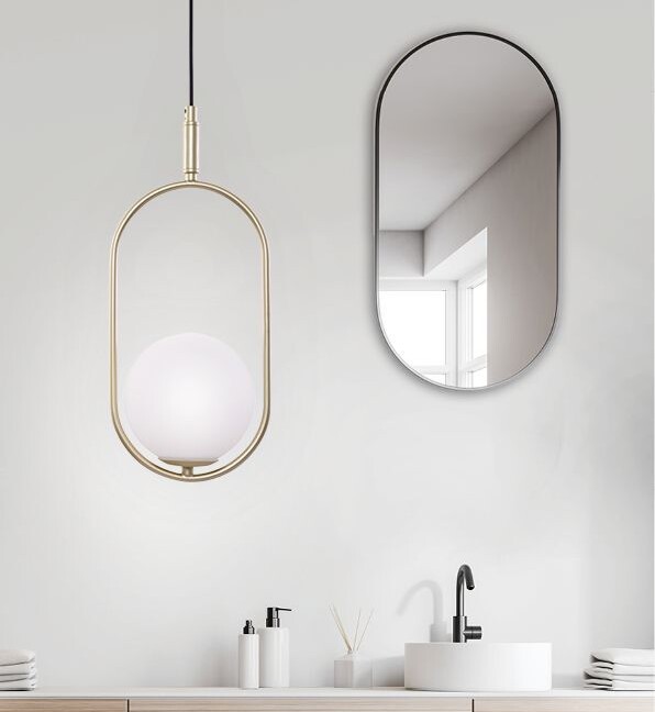 Złota lampa wisząca Cordel z białym kulistym kloszem wewnątrz owalnej, metalowej ramy, zawieszona w minimalistycznej łazience z owlanym lustrem, drewnianą szafką i białą umywalką nablatową.