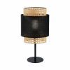Stylowa lampa stołowa Boho Nature z plecionym, rattanowym kloszem w naturalnym kolorze, uzupełnionym czarnym, tekstylnym pasem pośrodku. Osadzona na smukłej, metalowej nóżce z okrągłą podstawą. Ażurowa konstrukcja klosza tworzy ciekawy wzór światła i cienia. Idealna do wnętrz w stylu boho jako nastrojowe oświetlenie szafki nocnej lub komody.