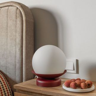 Nowoczesna lampa stołowa Kuul ST w kształcie kuli z matowego białego szkła osadzona na burgundowej, dwuczęściowej podstawie. Umieszczona na drewnianej szafce nocnej obok talerza z owocami liczi, przy tapicerowanym, beżowym zagłówku łóżka. Minimalistyczny design z kontrastującymi kolorami tworzy przytulną atmosferę w sypialni utrzymanej w naturalnych, ciepłych odcieniach.