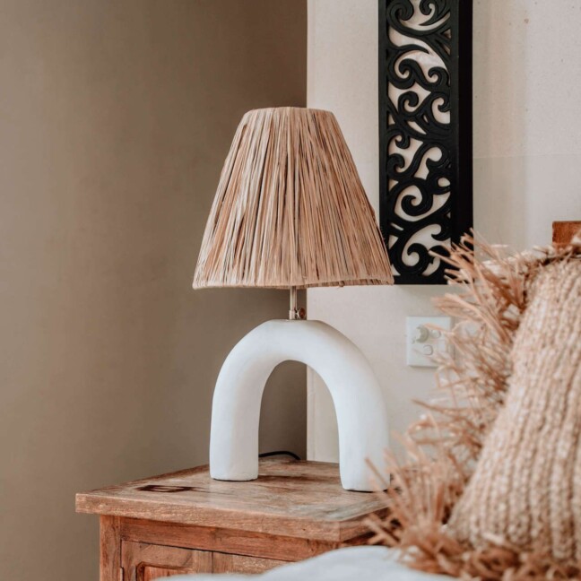 Designerska lampka nocna Mahalo S z charakterystyczną białą, łukową podstawą z ceramiki i naturalnym, beżowym abażurem z plecionych włókien. Ustawiona na drewnianym, rustykalnym stoliku nocnym w sypialni o neutralnych, ziemistych kolorach. Obok widoczna dekoracja z traw pampasowych, tworząca spójną, boho kompozycję idealną do przytulnych wnętrz w naturalistycznym stylu.