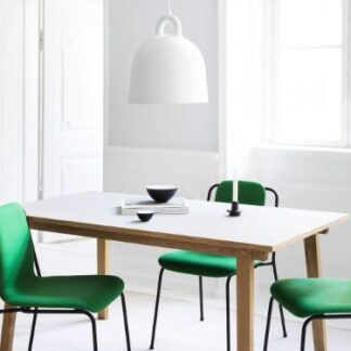 Biała lampa wisząca w kształcie dzwonu zawieszona nad minimalistycznym stołem z białym blatem i drewnianymi nogami. Jadalnia w skandynawskim stylu uzupełniona zielonymi krzesłami na ciemnych metalowych nóżkach. Jasne wnętrze z dużymi oknami zapewniającymi naturalne oświetlenie, podkreślające czystą, nowoczesną estetykę.