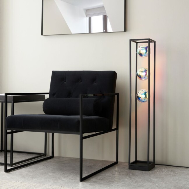 Nowoczesna lampa podłogowa Lumina z czarną metalową ramą w kształcie prostokąta i trzema złotymi, szklanymi kulami świetlnymi, ustawiona obok czarnego fotela w minimalistycznym, jasnym wnętrzu z lustrem na ścianie.