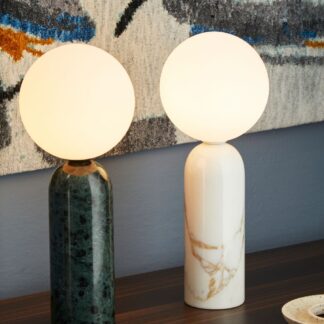 Dwie lampy stołowe z marmurową podstawą. Zielona i biała, z mlecznymi, kulistymi kloszami. Ustawione na drewnianym blacie na tle abstrakcyjnego dywanu. Elegancki, minimalistyczny design łączący naturalne kamienie z nowoczesną formą, idealny do współczesnych wnętrz.
