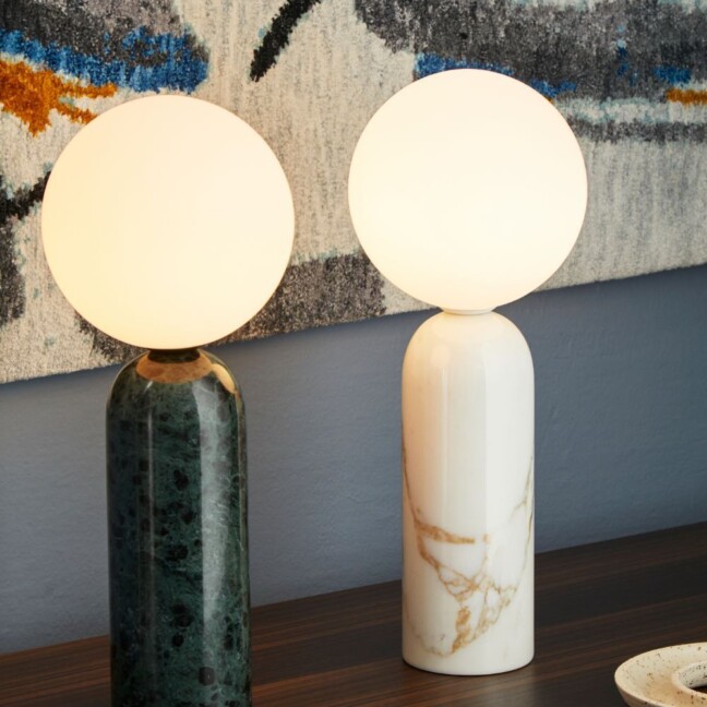 Dwie lampy stołowe z marmurową podstawą. Zielona i biała, z mlecznymi, kulistymi kloszami. Ustawione na drewnianym blacie na tle abstrakcyjnego dywanu. Elegancki, minimalistyczny design łączący naturalne kamienie z nowoczesną formą, idealny do współczesnych wnętrz.