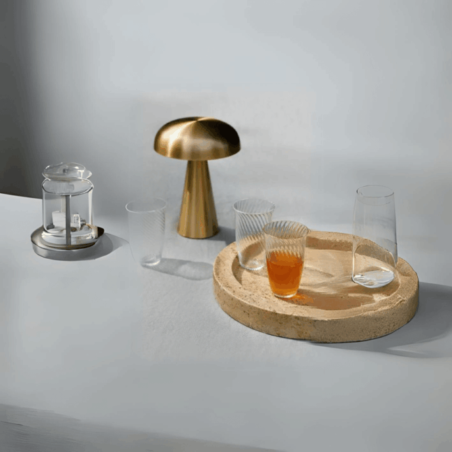 Przenośna lampa stołowa Como SC53 z mosiężnym, grzybkowym kloszem. Elegancki design z ciepłym, złotym wykończeniem, ładowana przez USB, ustawiona na jasnym stole.