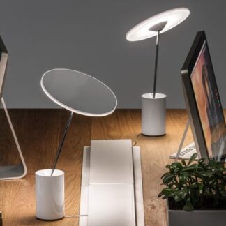 Nowoczesne lampki biurkowe Sisifo z okrągłym, ledowym talerzem na smukłym ramieniu i cylindryczną białą podstawą. Umieszczone na drewnianym biurku z komputerami, laptopami i akcesoriami do pracy. Minimalistyczny, regulowany design z dotykowym sterowaniem jasności, idealny do przestrzeni roboczych i domowych biur. Efektowne oświetlenie punktowe z możliwością precyzyjnego kierowania strumienia światła.