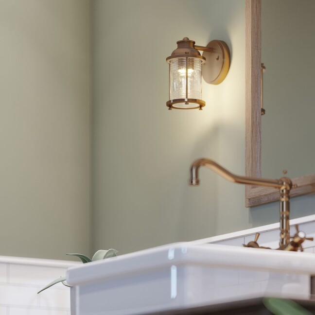 Mosiężny kinkiet Ashlandbay w stylu rustykalnym z latarniowym, szklanym kloszem. Elegancka lampa dworkowa w łazience z kremowymi ścianami i stylowym kranem. Ciepłe, przytulne oświetlenie do klasycznych wnętrz.