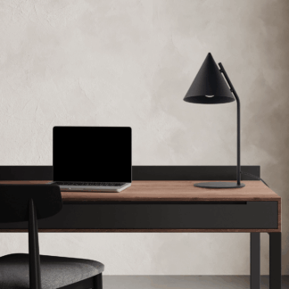 Minimalistyczna czarna lampa stołowa Cono TK w stylu japandi z charakterystycznym stożkowym kloszem na smukłej nóżce. Ustawiona na drewnianym biurku z czarną szufladą i metalowymi nogami, obok laptopa. Estetyka łącząca japońską prostotę i skandynawską funkcjonalność. Neutralne tło z teksturowaną, jasną ścianą podkreśla harmonijną kompozycję przestrzeni roboczej w stonowanych kolorach.
