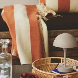 Minimalistyczna beżowa lampa stołowa w kształcie grzybka z półokrągłym kloszem, umieszczona na okrągłej, wiklinowej tacy. W tle widoczny koc w pomarańczowo-kremowe pasy na drewnianej ławie oraz szklana karafka. Ciepła, naturalna aranżacja w stylu boho z elementami rattanu, idealnie komponująca się z neutralnymi beżowymi odcieniami oświetlenia.