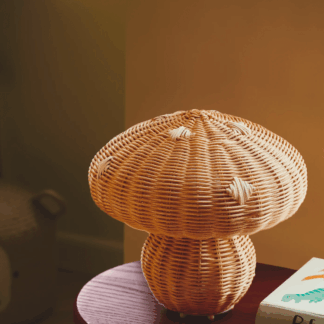 Rattanowa lampa stołowa Allie w kształcie grzyba, ręcznie pleciona z naturalnego materiału. Ustawiona na okrągłym stoliku na ciepłym, brązowym tle. Organiczna forma i naturalne wykończenie doskonale odzwierciedlają filozofię wabi sabi, celebrującą niedoskonałość.
