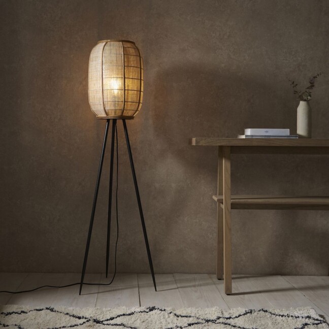 Lampa podłogowa Tanah z owalnym, rattanowym kloszem na czarnym trójnożnym stelażu, dająca ciepłe światło w przytulnym wnętrzu z brązową ścianą, drewnianym biurkiem i kremowym dywanem.
