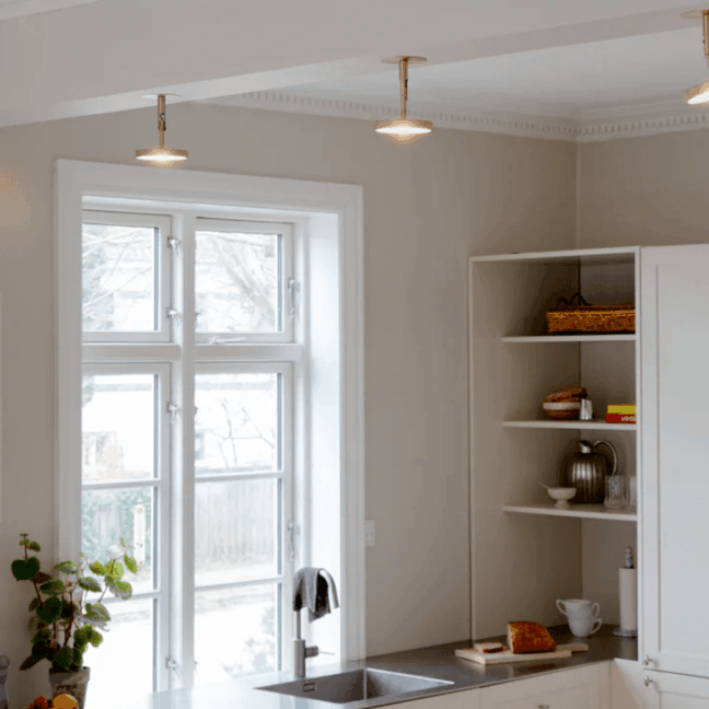 Złote lampy sufitowe LED Asteria Umage w mosiężnym wykończeniu nad białą kuchnią. Minimalistyczne reflektory równomiernie oświetlają przestrzeń roboczą z zlewozmywakiem. Eleganckie rozwiązanie do nowoczesnych wnętrz z klasycznymi elementami.