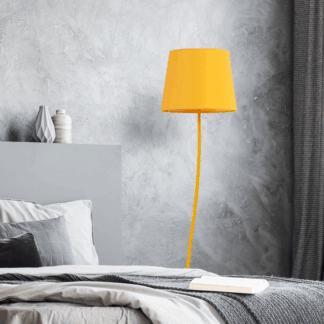 Żółta designerska lampa podłogowa Nicola z cytrynowym abażurem i dopasowaną podstawą, ustawiona przy szarej ścianie o teksturowanej powierzchni w nowoczesnej sypialni. W tle widoczne szare łóżko z białą i szarą pościelą oraz szare zasłony, tworząc minimalistyczną aranżację z kolorowym akcentem.