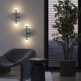 Nowoczesne kinkiety LED z dotykowym włącznikiem na białej ścianie. Lampy z kulistymi kloszami na pionowych ramionach tworzące nastrojowe oświetlenie w eleganckim, minimalistycznym salonie z szarymi meblami.