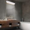 Nowoczesna biała lampa ścienna nad lustrem w łazience. Minimalistyczny kinkiet łazienkowy o podłużnym kształcie emitujący jasne światło, zamontowany w ciemnej, eleganckiej aranżacji z drewnianą szafką i wolnostojącą wanną.