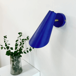 Niebieski kinkiet Cale w minimalistycznym stylu z regulowanym, stożkowym kloszem i włącznikiem. Nowoczesna lampa ścienna na białej ścianie obok wazonu z zielenią. Designerskie oświetlenie punktowe w intensywnym kobaltowym kolorze.