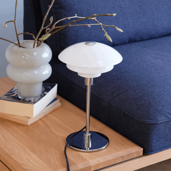 Elegancka lampa stołowa Morph z charakterystycznym mlecznym, trójwarstwowym szklanym kloszem w kształcie grzybka i chromowaną, okrągłą podstawą. Umieszczona na drewnianym stoliku obok granatowej sofy, białego ceramicznego wazonu z gałązkami i stosu książek. Nowoczesny, skandynawski design z neutralną kolorystyką zapewniający przyjemne, rozproszone światło, idealne do strefy wypoczynkowej salonu.