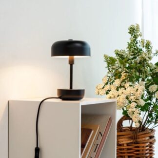 Czarna lampka stołowa Haipot w kształcie grzybka z płaskim, okrągłym kloszem na smukłej nóżce i cylindrycznej podstawie. Umieszczona na białej szafce z książkami, obok wiklinowego kosza z kwitnącymi białymi roślinami. Minimalistyczny, nowoczesny design z matowym wykończeniem doskonale komponuje się z jasnym wnętrzem, zapewniając stylowe oświetlenie punktowe do czytania.
