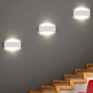 Trzy białe nowoczesne kinkiety LED Concha zamontowane na szarej ścianie wzdłuż drewnianych schodów. Płaskie, prostokątne oprawy emitują ciepłe światło górą i dołem, tworząc dekoracyjny efekt świetlny oraz zapewniając oświetlenie klatki schodowej.