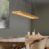 Drewniana liniowa lampa wisząca Brad LED zawieszona nad dużym, masywnym stołem z litego drewna, otoczonym jasnymi, nowoczesnymi krzesłami. W tle widoczna szara ściana z abstrakcyjnym obrazem w zielonych tonach oraz drewniana podstawka ze świecami na stole.