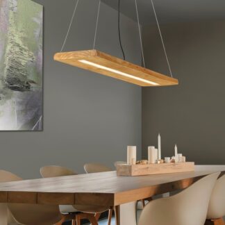 Drewniana liniowa lampa wisząca Brad LED zawieszona nad dużym, masywnym stołem z litego drewna, otoczonym jasnymi, nowoczesnymi krzesłami. W tle widoczna szara ściana z abstrakcyjnym obrazem w zielonych tonach oraz drewniana podstawka ze świecami na stole.