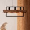 Loftowa lampa wisząca Solvane 3 - połączeniem czarnej metalowej ramy, drewnianej belki i trzech żarówek typu Edison. Zawieszona na tle białej ściany i drewnianego panelu, uzupełniona dekoracyjną trawą pampasową u dołu.